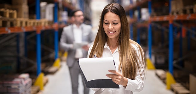 Das Foto zeigt eine Frau mit einem Klemmtbrett in einer Lagerhalle.